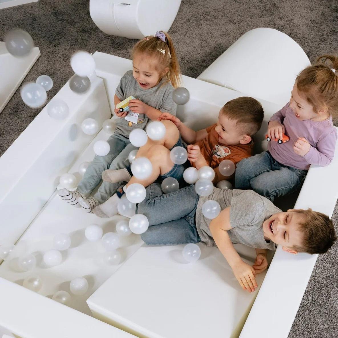 Four children sitting inside white foam ball pit filled with white and gray sensory play balls