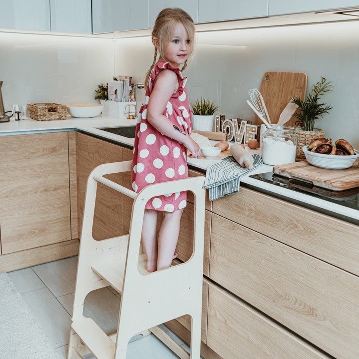 Girl in pink polka dot dress standing on white kitchen helper tower step stool