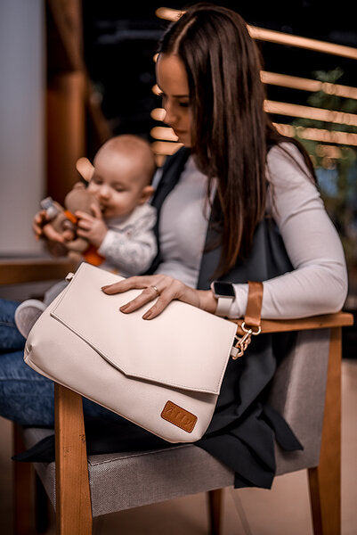 Mother holding sleeping infant and cappuccino handbag across body with long shoulder strap
