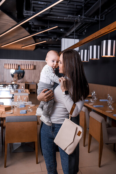 Woman holding baby with cappuccino mum bag worn on shoulder in contemporary space