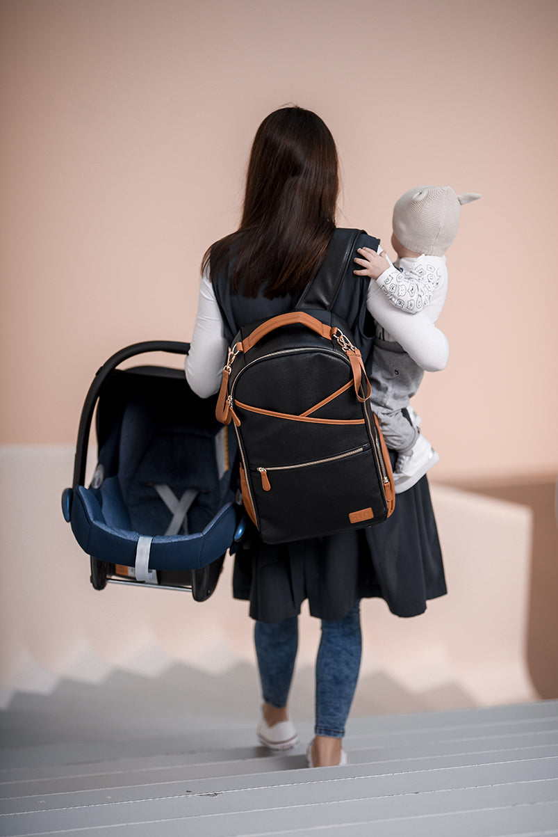 Woman holding toddler wearing black coffee diaper backpack with infant car seat