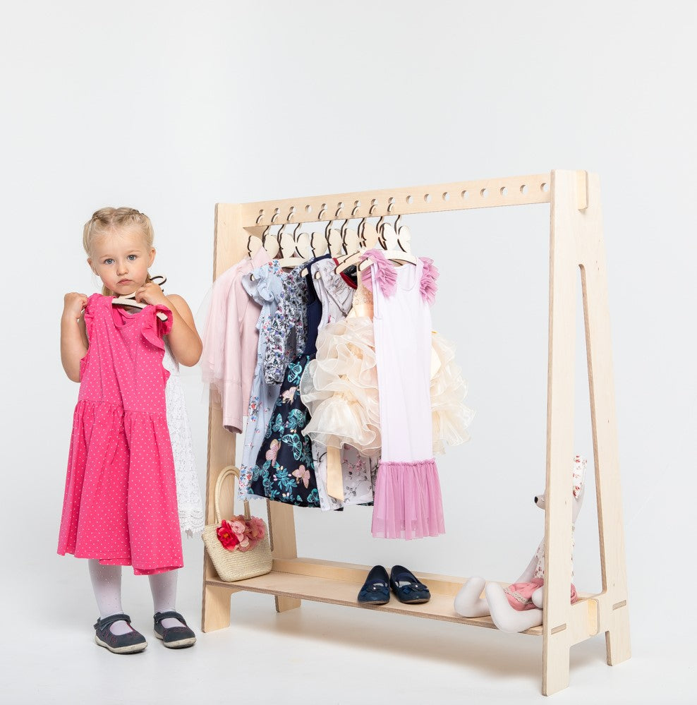 Young toddler girl holding pink dress while standing next to wooden clothing rack