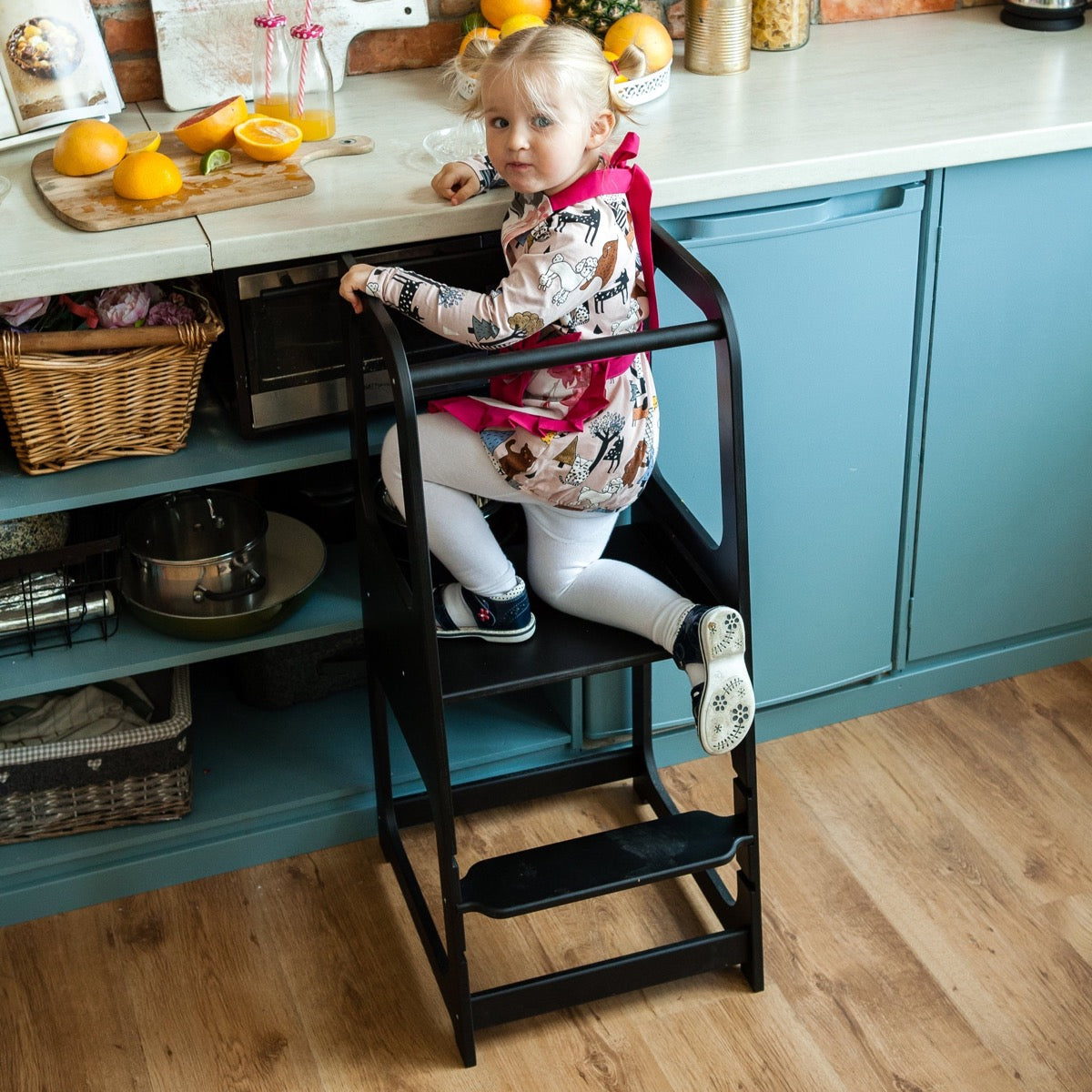 Toddler standing on gray helper tower stool preparing food at kitchen counter