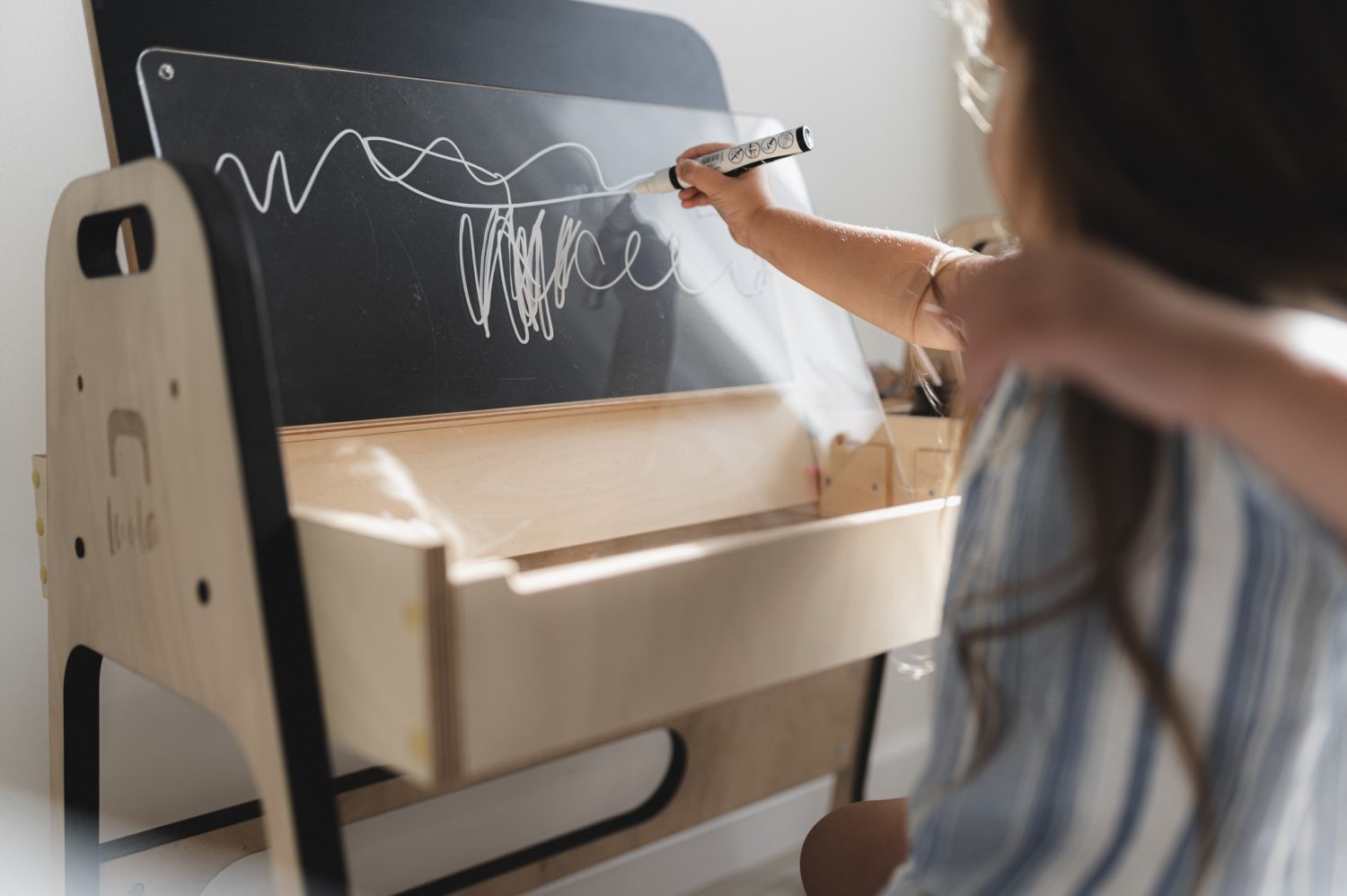 Child writing on chalkboard panel while sitting at Montessori table with storage