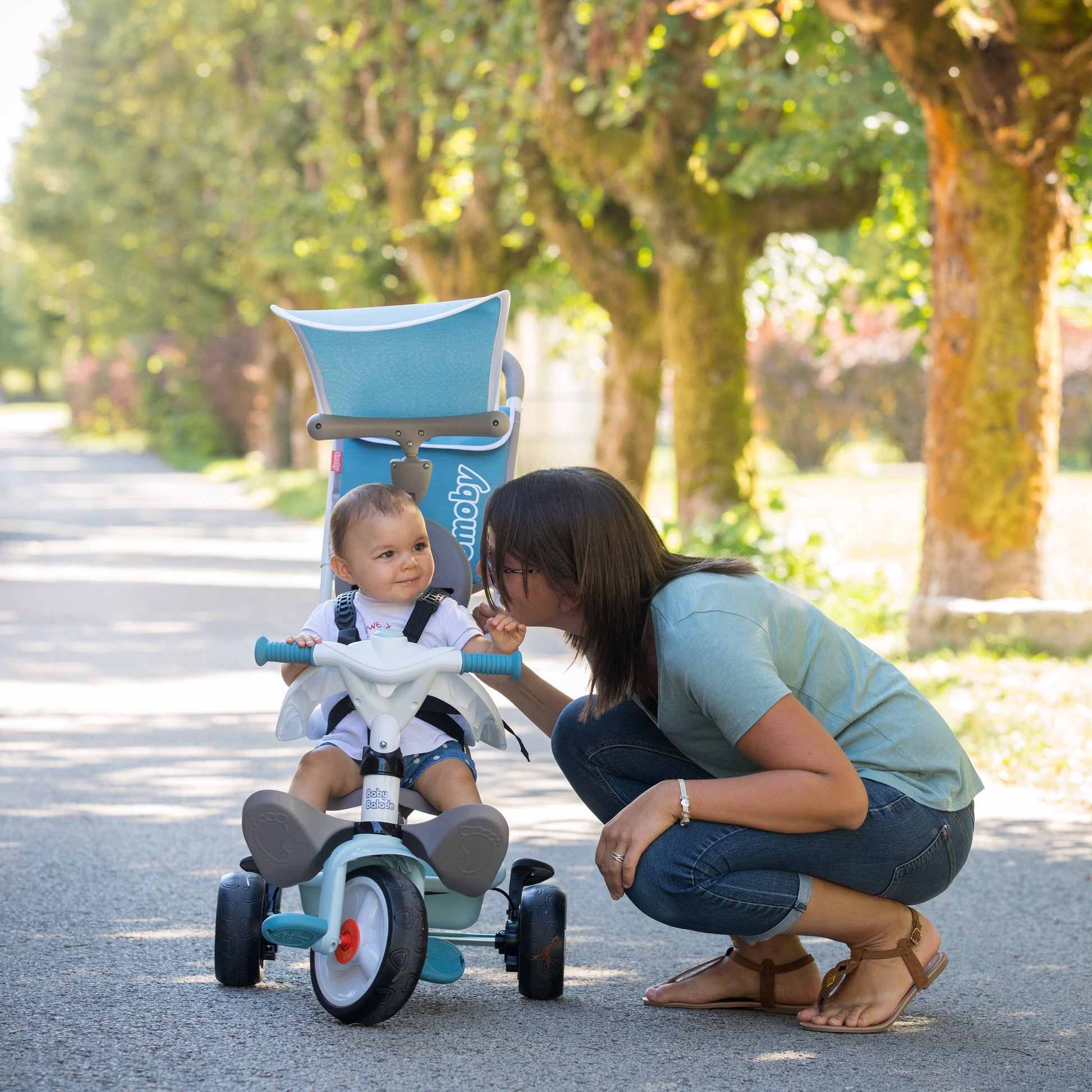 Toddler riding blue Baby Balade tricycle outdoors while parent supervises on tree-lined path