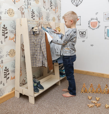 Toddler boy selecting clothes from natural wood Montessori dress-up rack with mirror