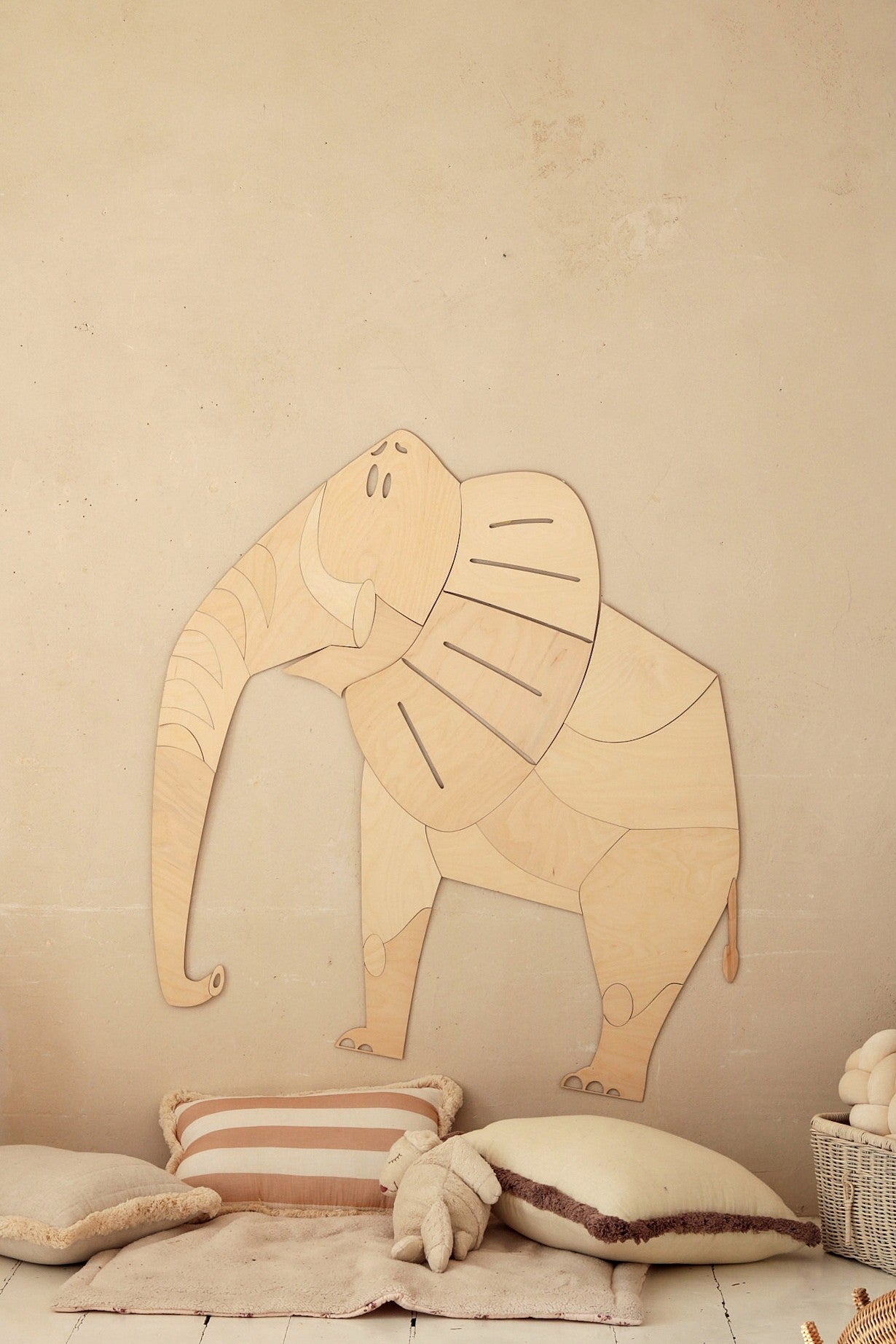 Wooden elephant wall decoration mounted on beige wall above cushions and toys in nursery