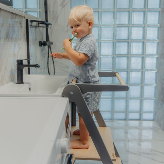 Toddler in grey shirt reaching countertop while standing on natural wood kitchen tower with safety features