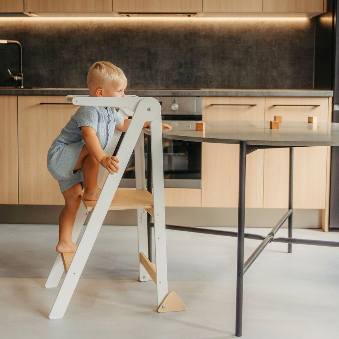 Child in blue shirt on white kitchen tower at counter height observing cooking activity at modern stove