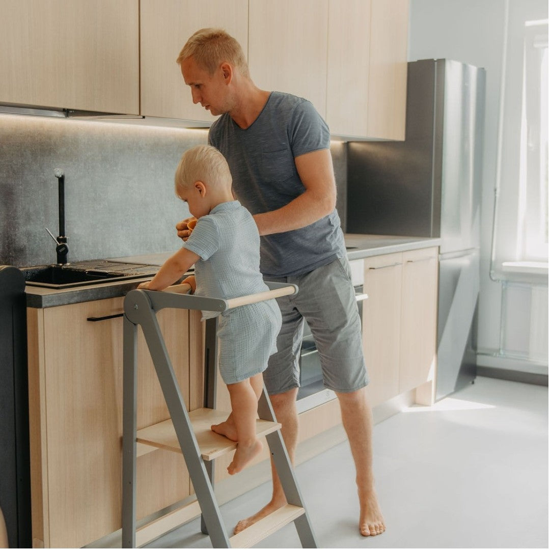 Adult supervising toddler on grey kitchen tower at counter while child stands at raised platform cooking