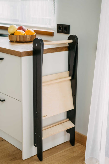 Black foldable kitchen tower standing closed against white kitchen cabinet with removable rails stored inside