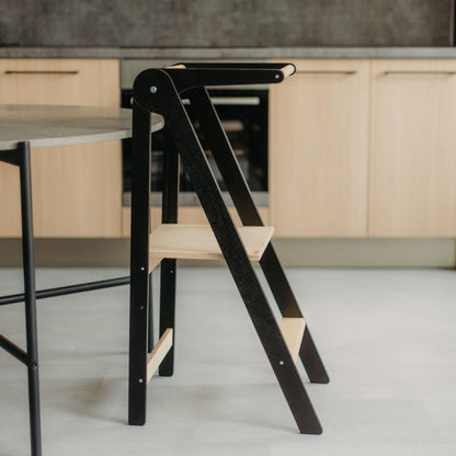 Young child on natural wood kitchen tower with removable rail at oven height in modern kitchen setting