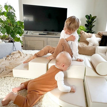 Two toddlers playing and climbing on assembled beige foam block structures in living room