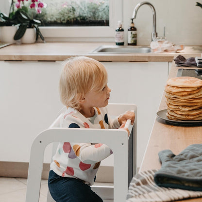 Toddler using white Montessori kitchen tower to reach counter and prepare pancakes safely