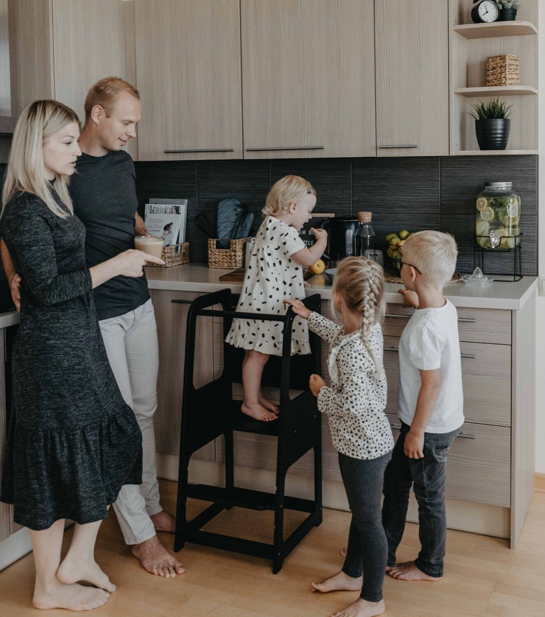 Family using black kitchen helper tower together, children standing at counter height to cook