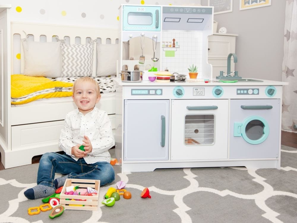 Toddler playing with wooden kitchen set in bedroom with accessories and toy food scattered around