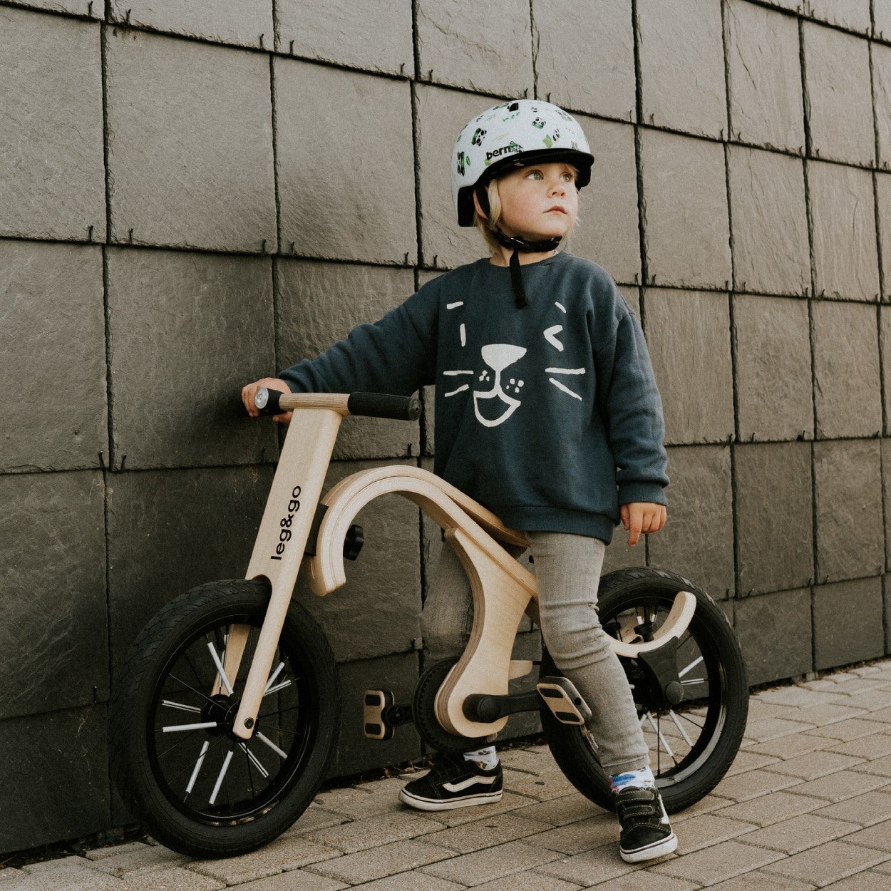 Toddler standing with leg&go pedal bike against textured wall backdrop