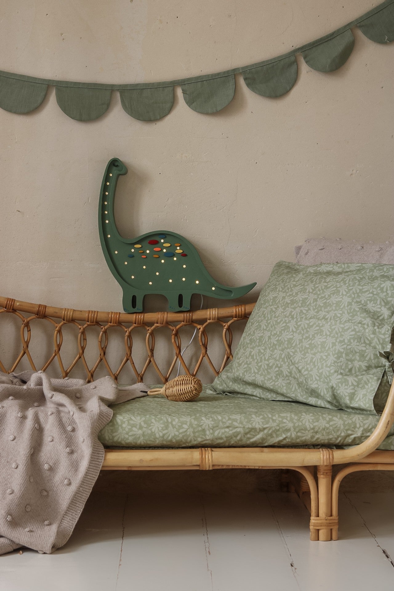 Green dinosaur lamp mounted above woven bed with green bedding and draped wall garland in nursery