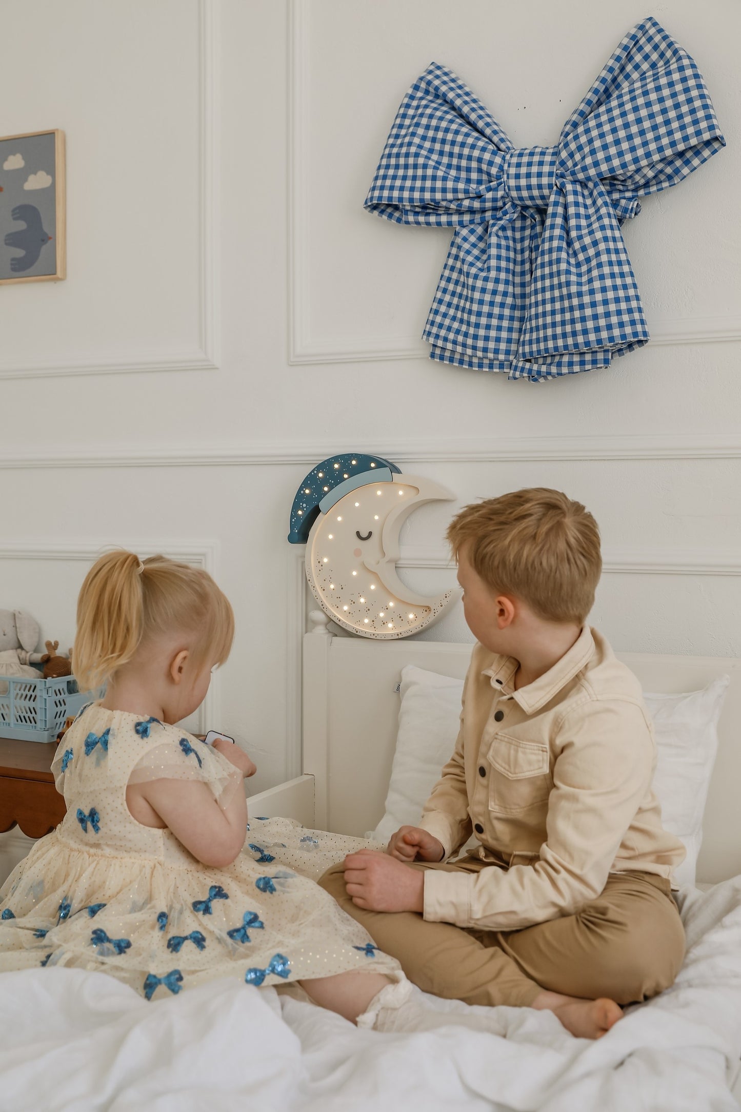 Two young children sitting on bed looking at illuminated moon lamp hanging on wall