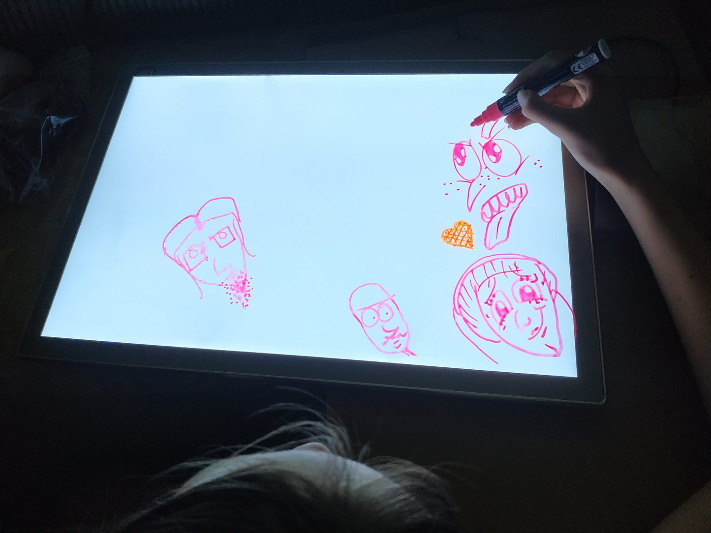 Close-up of child drawing pink and orange sketches on lit light board