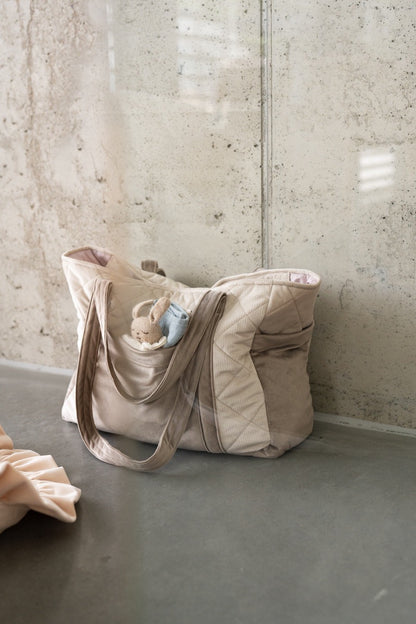 Beige mom weekender bag holding folded baby clothes and items, angled side view indoors