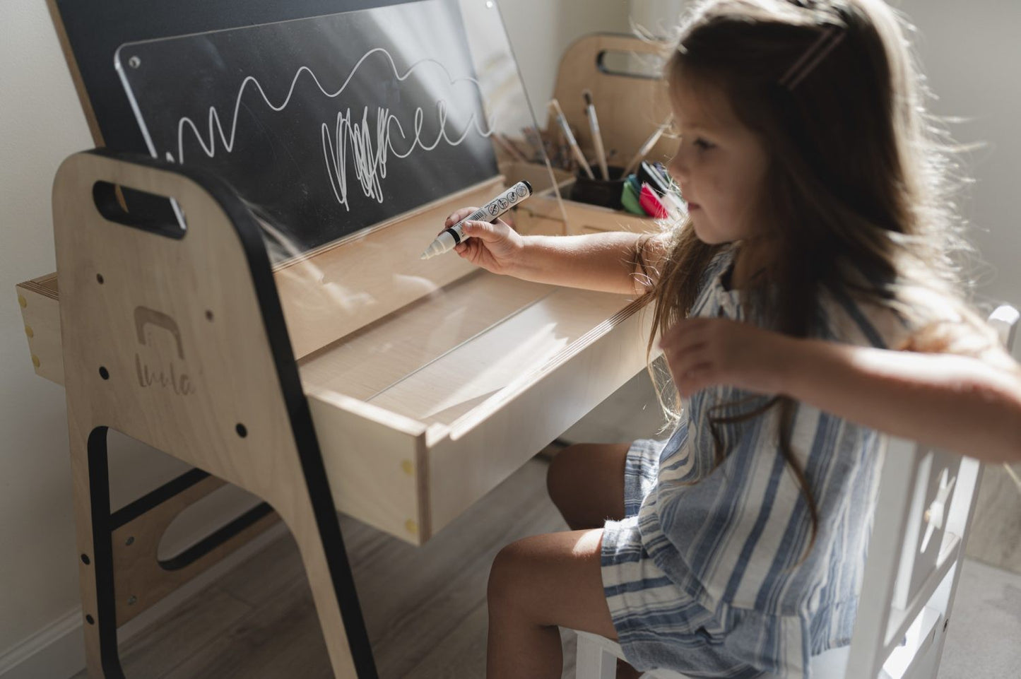Child writing on whiteboard surface of Julle desk with art supplies organized nearby