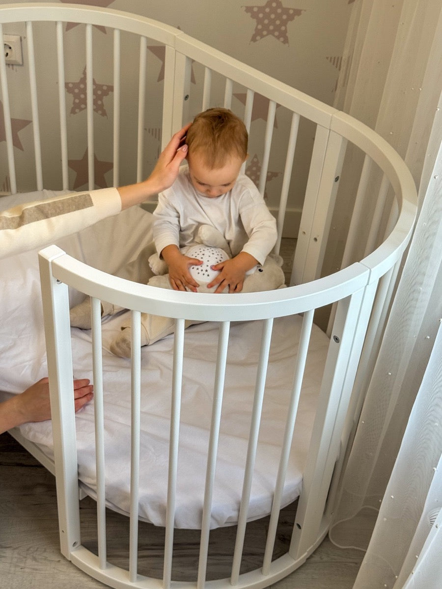 Overhead view of smiling baby sleeping in white round crib with neutral bedding and blanket