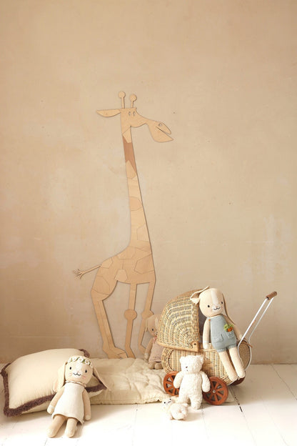 Wooden giraffe wall art standing next to wicker chair with stuffed animals and toys on white floor