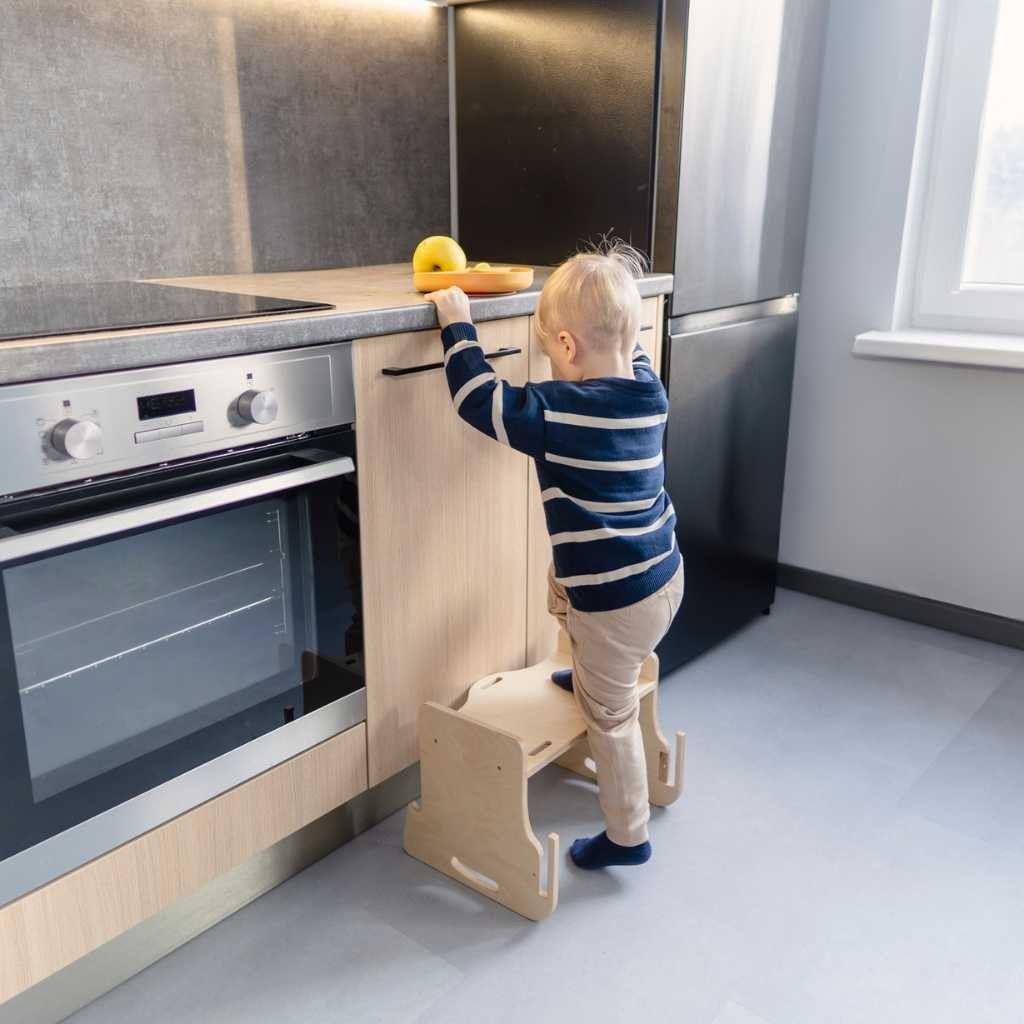 Child reaching up to stovetop while standing on wooden kitchen learning tower