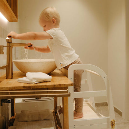 Young child reaching toward water bowl while standing in white kitchen tower