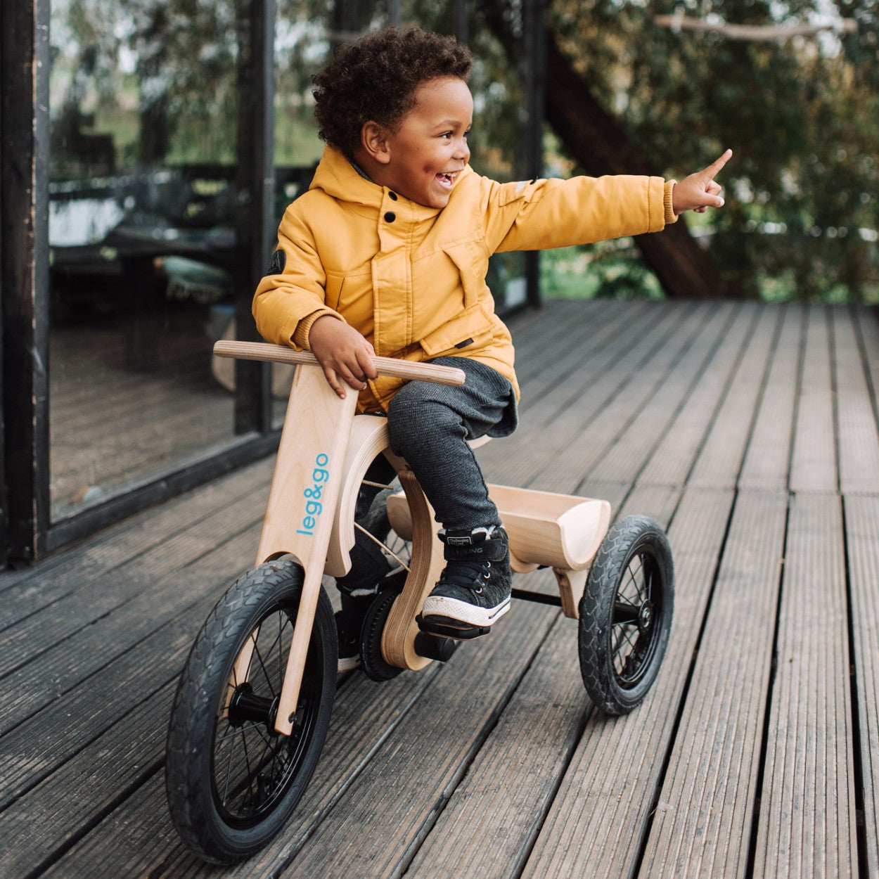 Smiling child in yellow jacket riding tricycle add-on on wooden deck