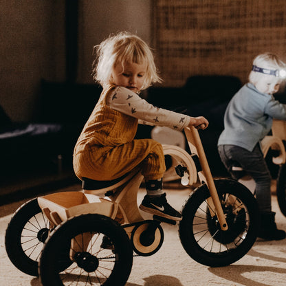 Toddler steering wooden tricycle add-on indoors with another child nearby