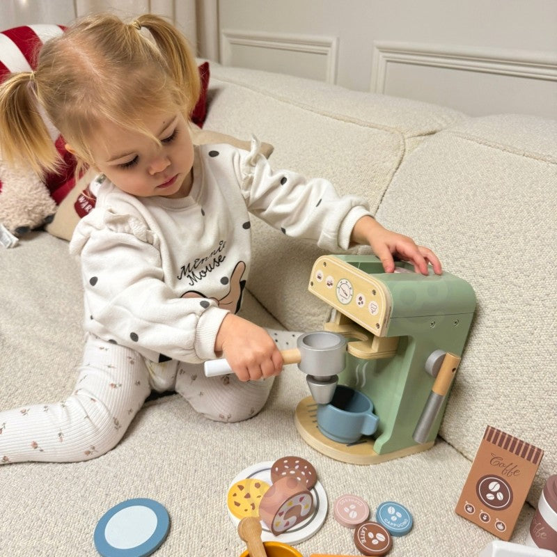 Toddler reaching toward wooden coffee machine while seated on couch with accessory pieces nearby